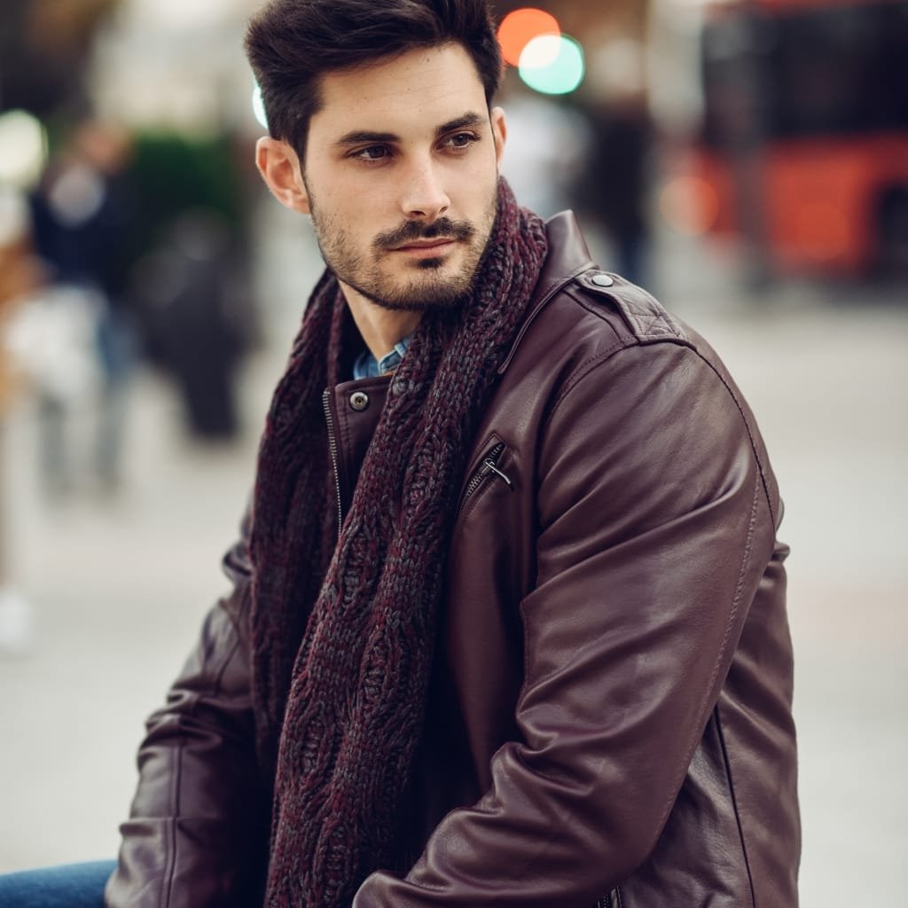 A stylish man with short dark hair and a neatly trimmed beard sits outdoors. He wears a brown leather jacket and a chunky maroon scarf, looking off to the side with a thoughtful expression. The background is a softly blurred urban street.