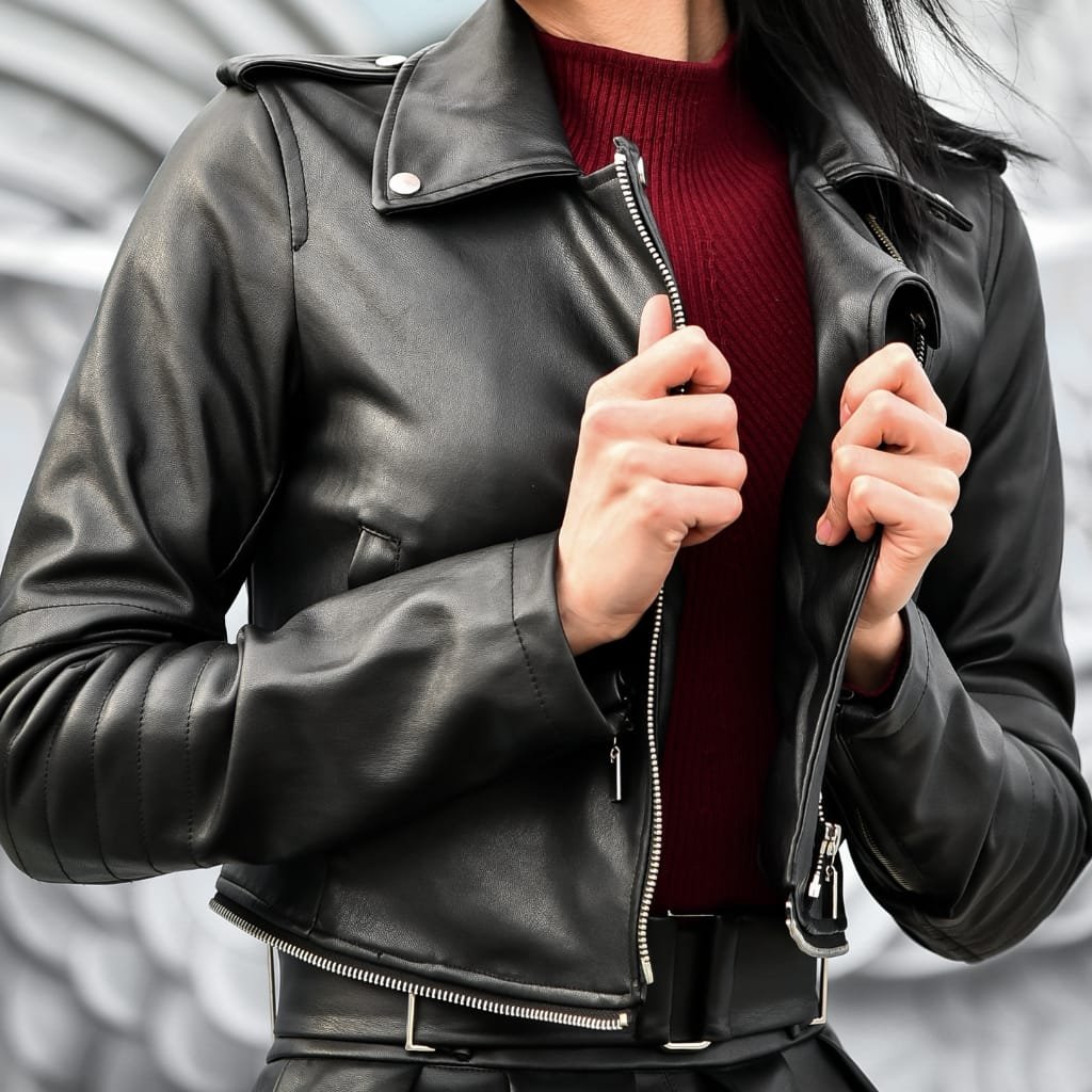 A close-up of a woman wearing a black leather motorcycle jacket, zipping up the front. She is also wearing a burgundy knit sweater underneath. The jacket has quilted elbow panels and silver hardware.
