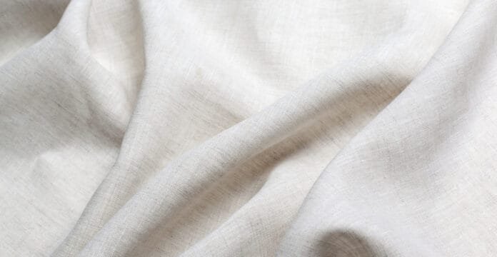 A close-up of soft, light-colored linen blend fabric with gentle folds and natural weave texture.