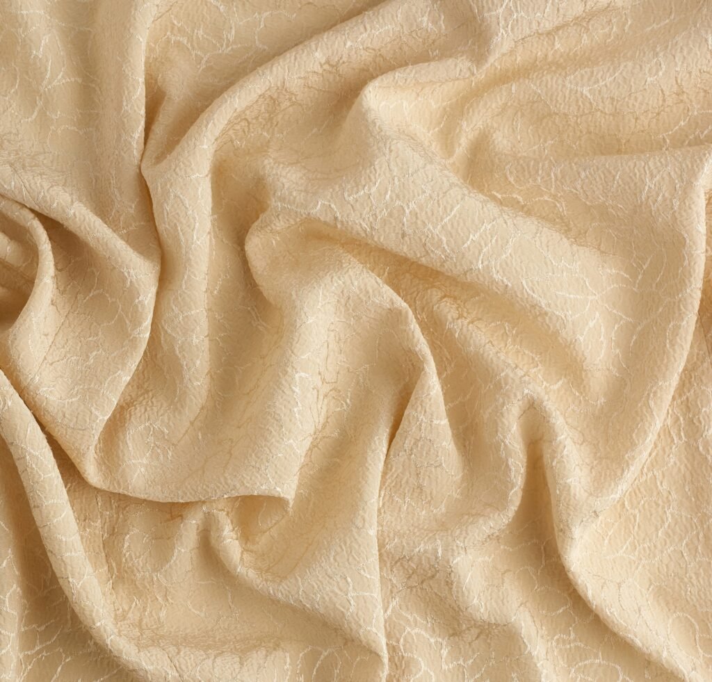 A close-up photograph of beige linen crepe fabric with a soft, crinkled texture and delicate, irregular veining. The fabric forms graceful folds and shadows, highlighting its lightweight, airy drape.
