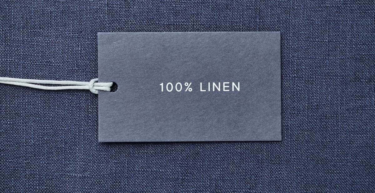 "Dark blue linen fabric background with a small gray certification tag reading '100% LINEN' and white string tied on the left."