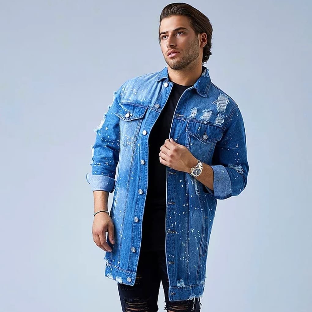 A man wearing a long, distressed blue denim jacket over a black shirt, with the sleeves rolled up and a silver wristwatch, posing against a light blue background.