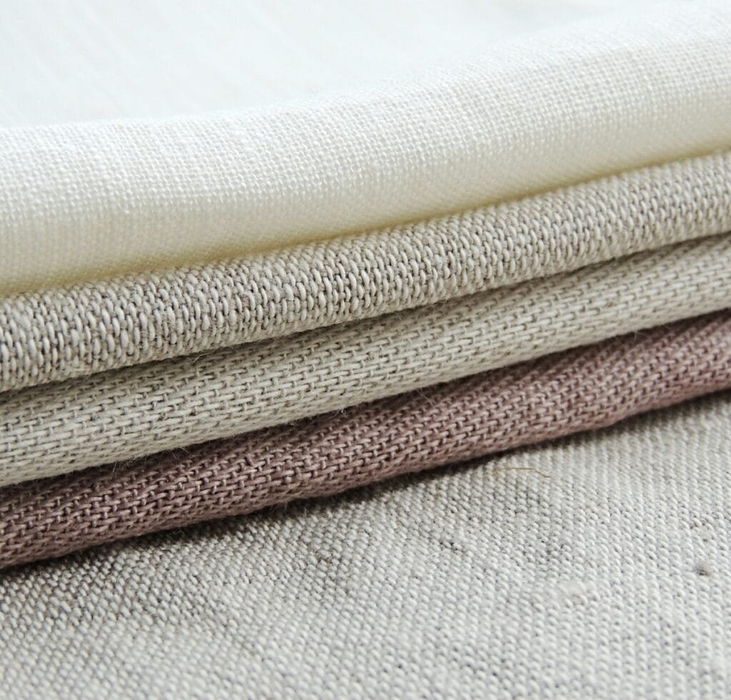 A close-up stack of loosely woven linen fabrics in soft, natural earth tones including ivory, light beige, taupe, and dusty pink. The textiles show pronounced, airy weaves with visible irregularities and a textured, breathable surface.