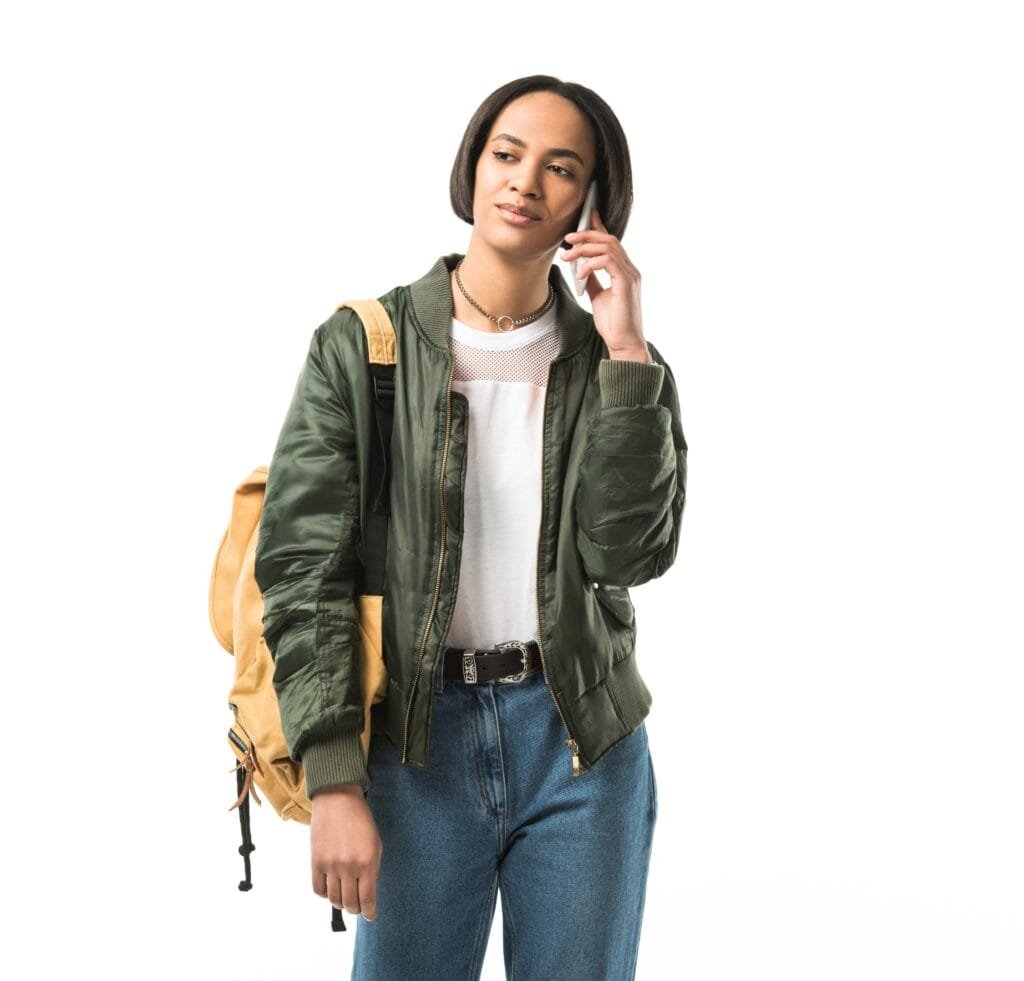 “Woman wearing a green MA-1 bomber jacket with a white mesh top, blue jeans, and a tan backpack, talking on a cellphone.”
