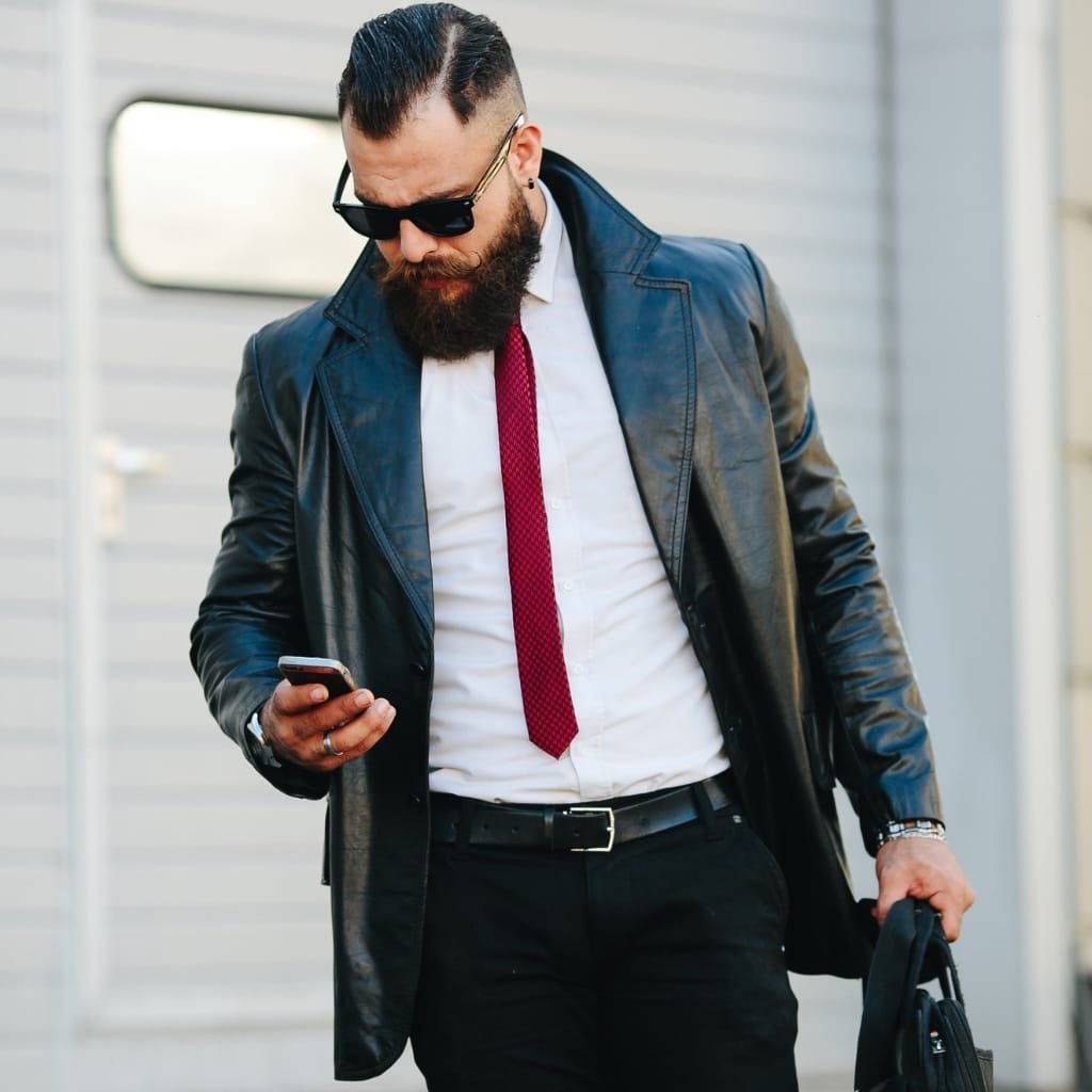 "A stylish man wearing a blazer-style leather jacket, white dress shirt, red tie, black pants, and sunglasses. He has a full beard and is looking at his phone while carrying a bag."