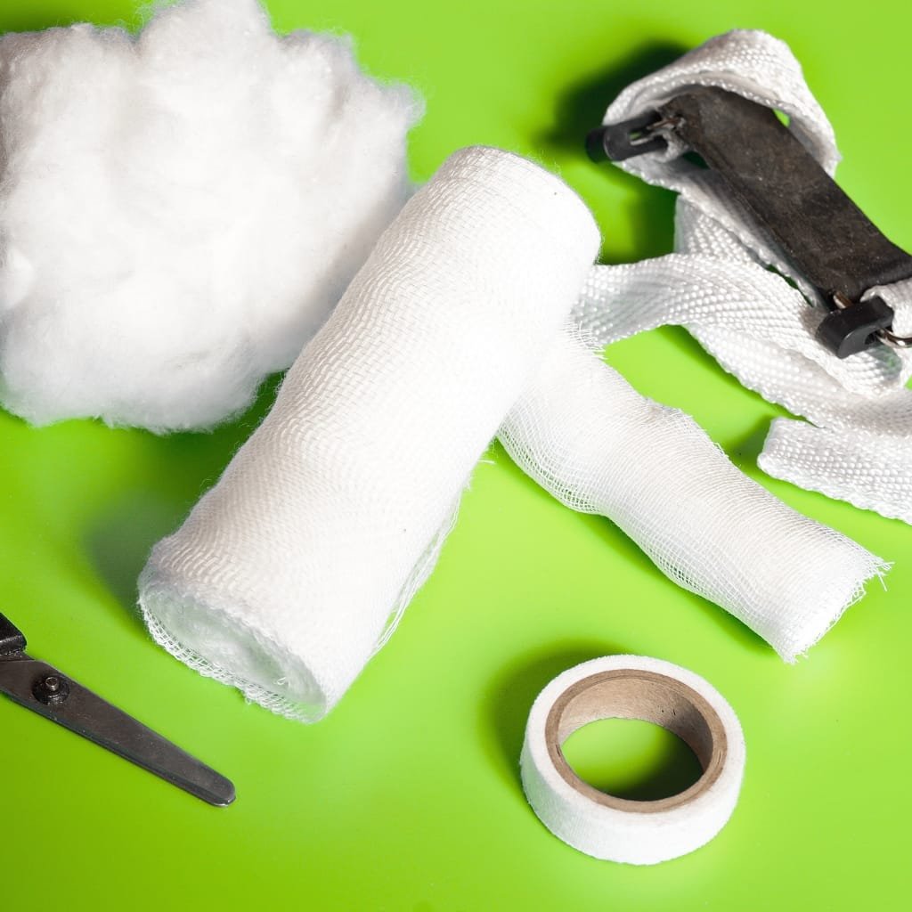 A collection of medical cotton items on a bright green background: a roll of gauze, a fluffy cotton ball, a stripped bandage fabric, a metal clamp or tensioner, and a roll of medical tape.