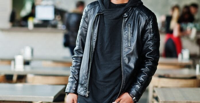 A man wearing a black blazer-style leather jacket and a black shirt, standing in a casual indoor setting.