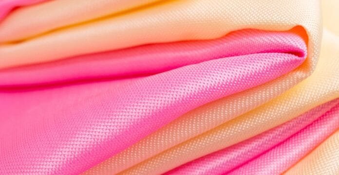 A close-up of folded moisture-wicking polyester fabric in varying pink and peach tones, showing a smooth, woven texture.