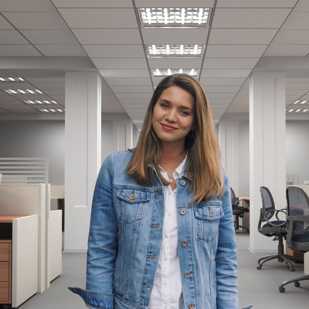 Young woman in a denim jacket standing in a modern open-plan office with cubicles and desks in the background.