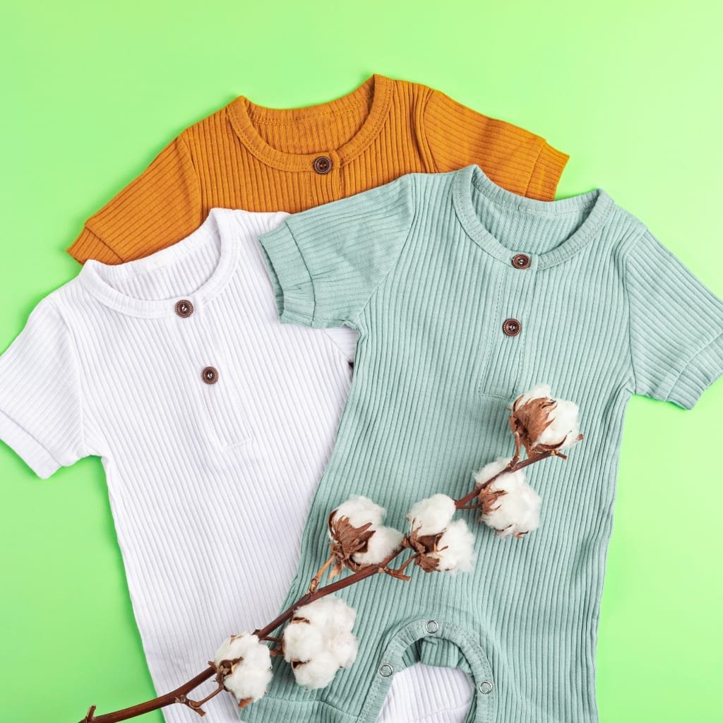 Close-up of three ribbed organic cotton baby onesies in white, orange, and mint green, arranged on a bright green background with a cotton branch.