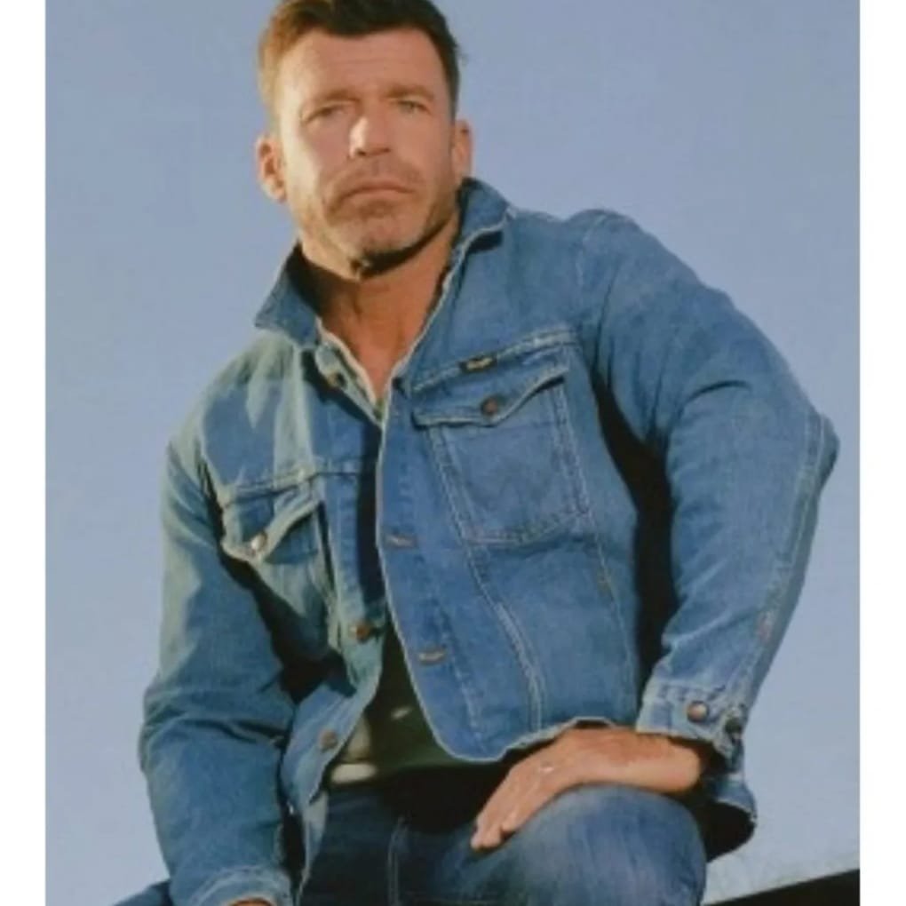 "Man wearing a blue denim jacket, seated outdoors against a clear sky, with a serious expression and short dark hair."