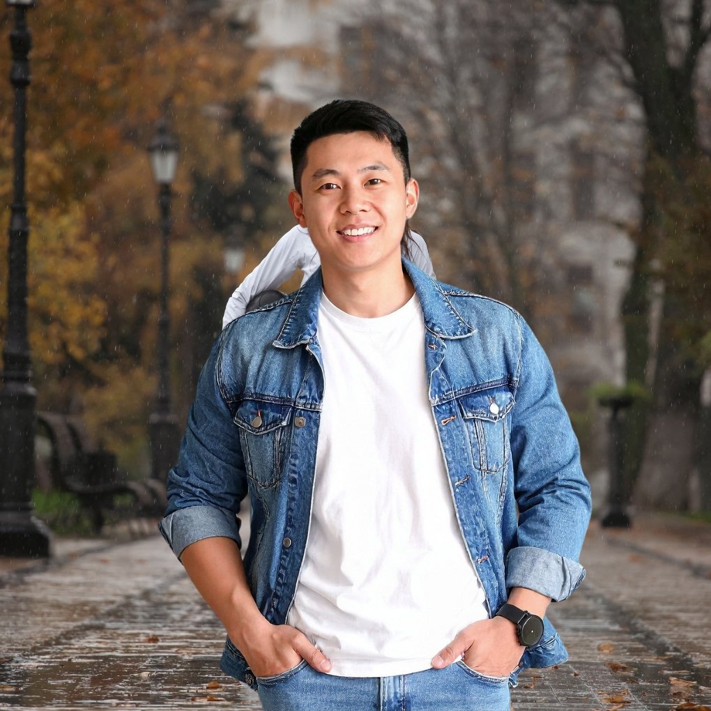 Young man wearing a blue denim jacket over a white t-shirt, outdoors on a rainy autumn day with trees and lampposts in the background.