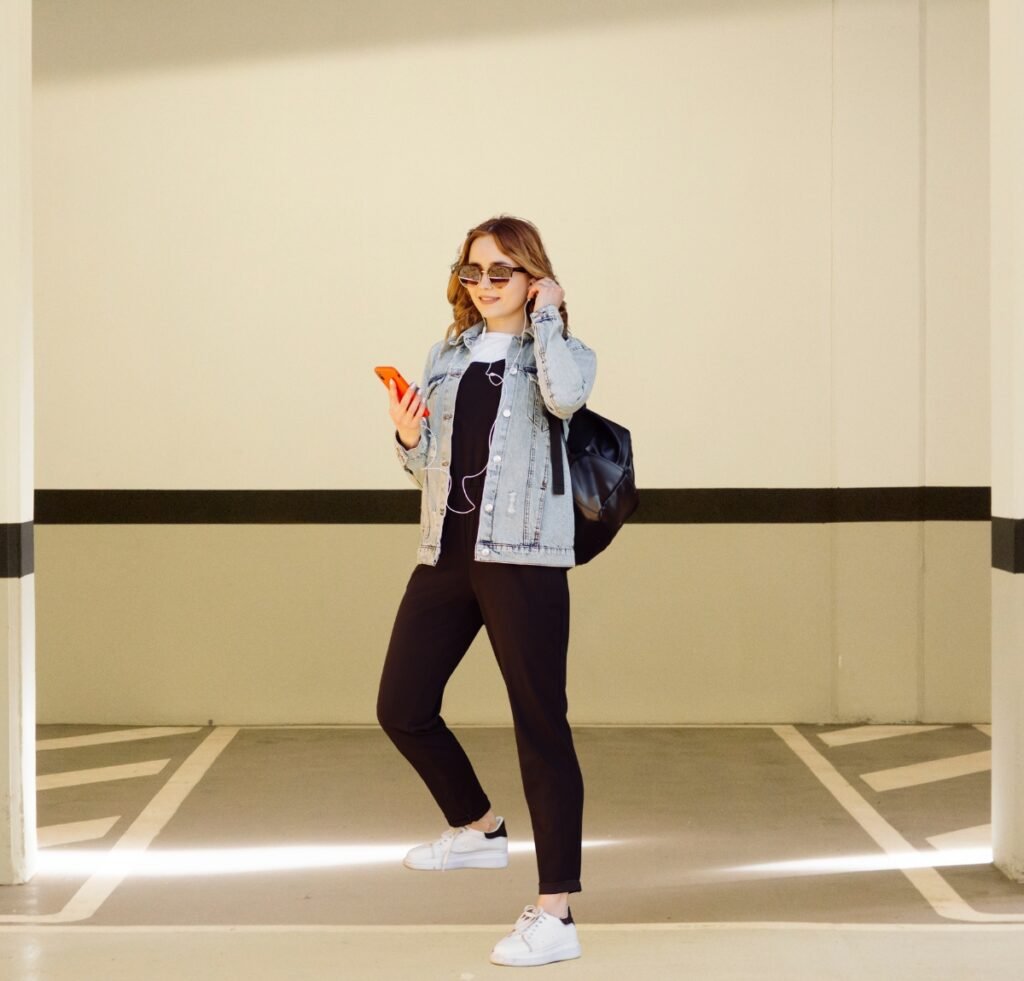 A stylish young woman stands in a parking garage wearing a casual denim bomber jacket over a black top, black pants, white sneakers, and sunglasses. She holds an orange phone in one hand and has a black backpack slung over one shoulder.