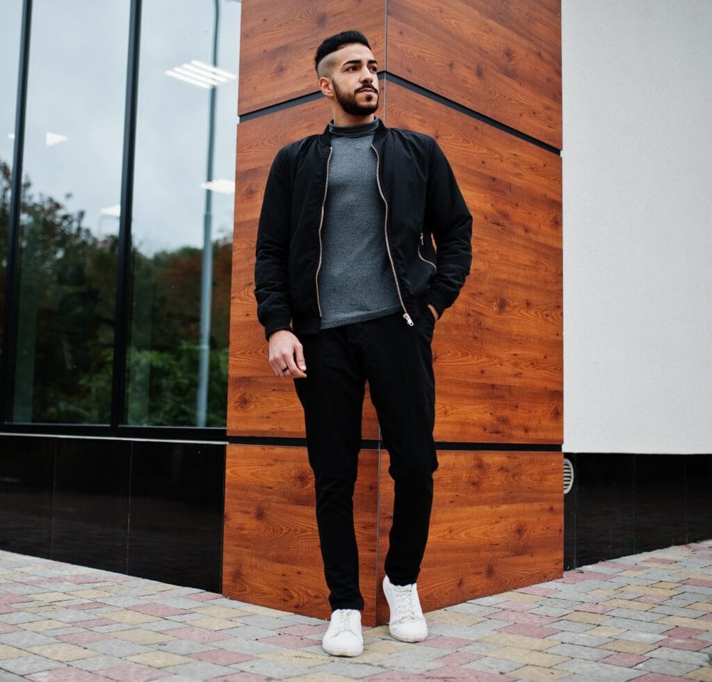 A man stands casually outdoors against a modern wooden panel wall and glass storefront. He wears a black bomber jacket over a gray turtleneck or knit sweater, black slim pants, and white sneakers. The overall vibe is sleek and contemporary, blending casual ease with a polished edge.
