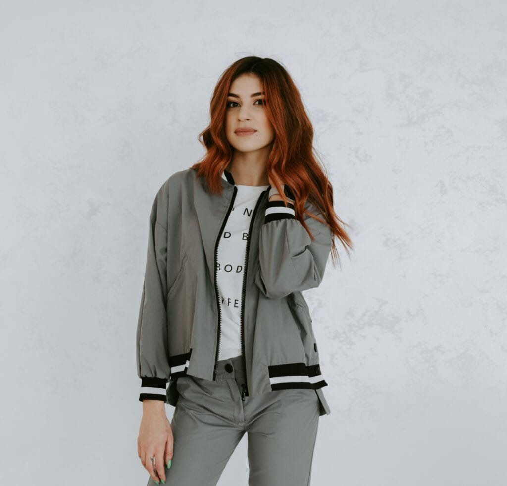 "Woman wearing a light gray bomber jacket over a graphic white T-shirt and gray pants, posing against a light textured wall."