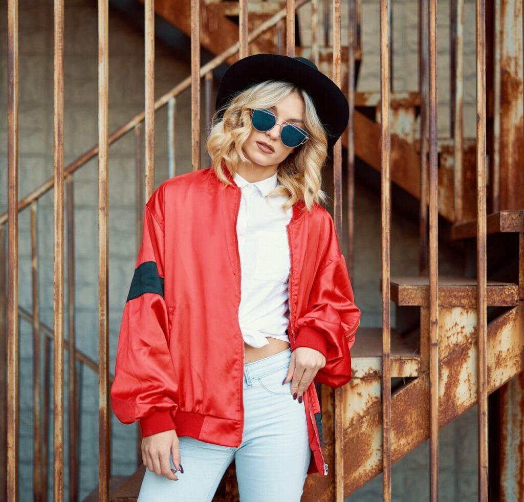A stylish young woman wearing an oversized red bomber jacket with a black cuff patch, light-washed jeans, a white button-up shirt tied at the waist, a black wide-brim hat, and blue-tinted sunglasses. She stands beside rusted metal railings and stairs in an urban setting. Types of Bomber Jackets