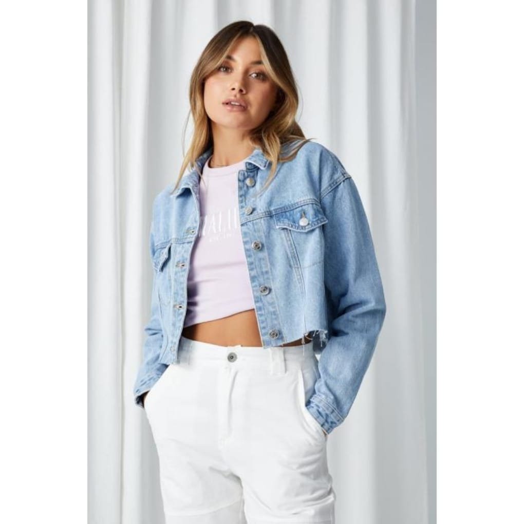 A young woman wearing a light wash denim jacket over a pale pink cropped top and white high-waisted pants, standing with hands in pockets in front of a light curtain backdrop.