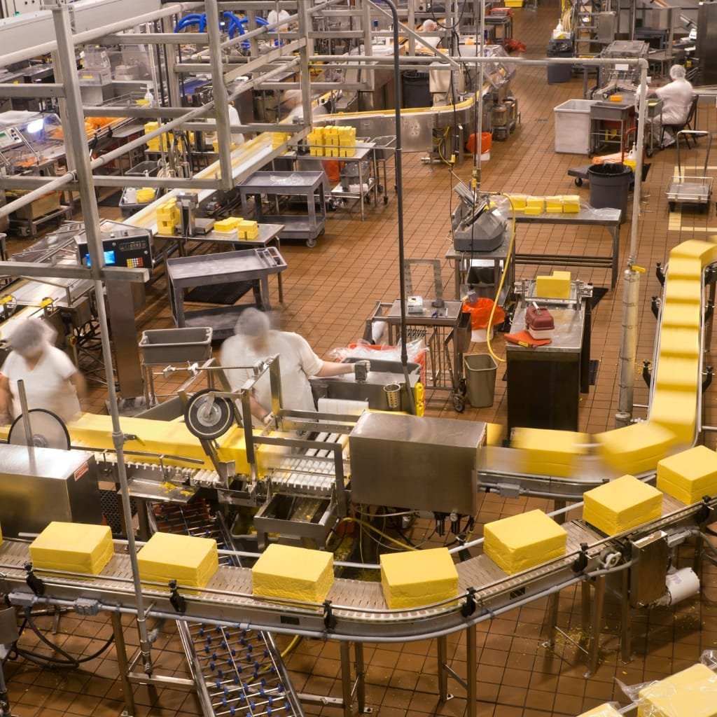 A busy industrial food processing line with yellow blocks moving along a conveyor belt; workers in white uniforms and hairnets operate equipment amidst stainless steel machinery and orange-brown tiled floors.