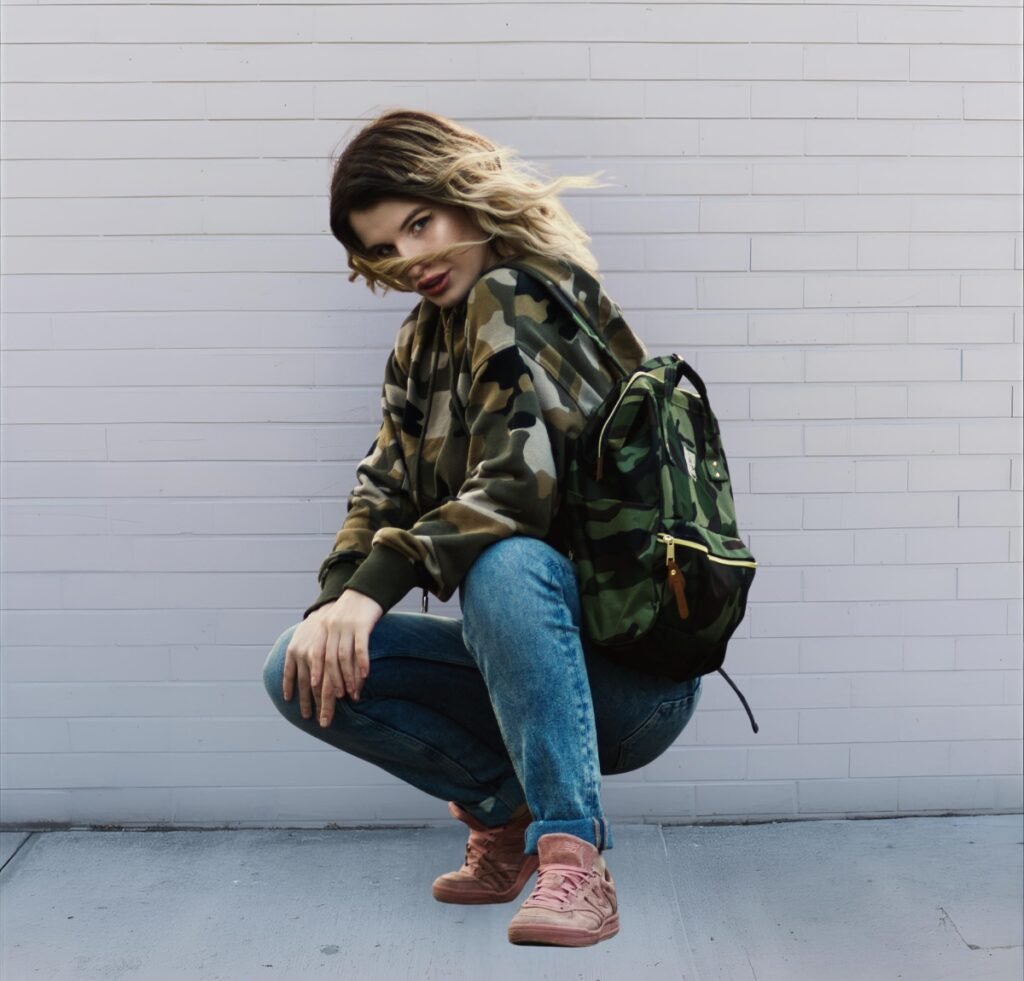 A stylish young woman crouches against a light gray brick wall, wearing a camouflage bomber jacket, blue jeans, pink sneakers, and a matching camouflage backpack.