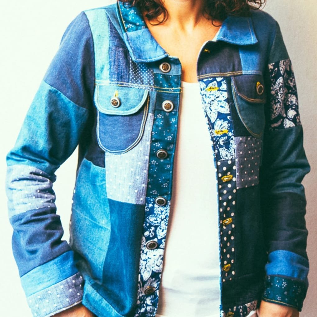 A person wearing a patchwork denim jacket in various blue tones, with a light-colored inner top visible, standing against a neutral background.