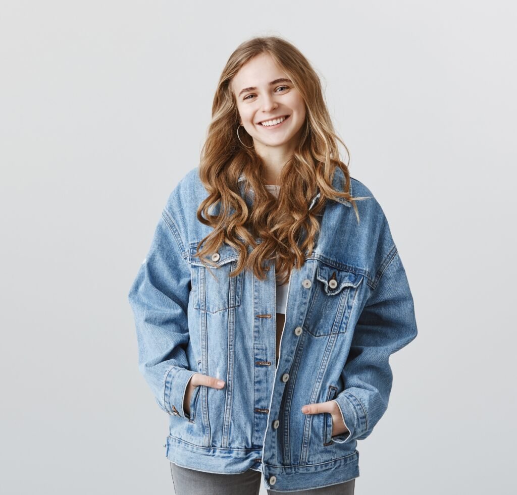 A smiling young woman stands against a light gray background, wearing an oversized light blue denim jacket over a white top. Her long wavy hair frames her face, and she has her hands in the jacket pockets.