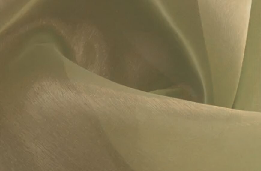 A close-up view of lightweight, sheer Polyester Organza fabric with a soft, pale gold/cream color. The fabric drapes in gentle folds, showing a subtle, silky sheen and a fine, crisp texture that catches light along the smooth surface and edges. polyester organza fabric