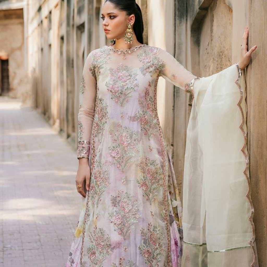 A model wearing a pastel pink and lavender floral embroidery long dress with sheer organza sleeves and a matching dupatta in a soft, airy fabric. She stands in a sunlit alley, accessorized with statement earrings and bracelets, showcasing delicate embellishments and scalloped borders.
