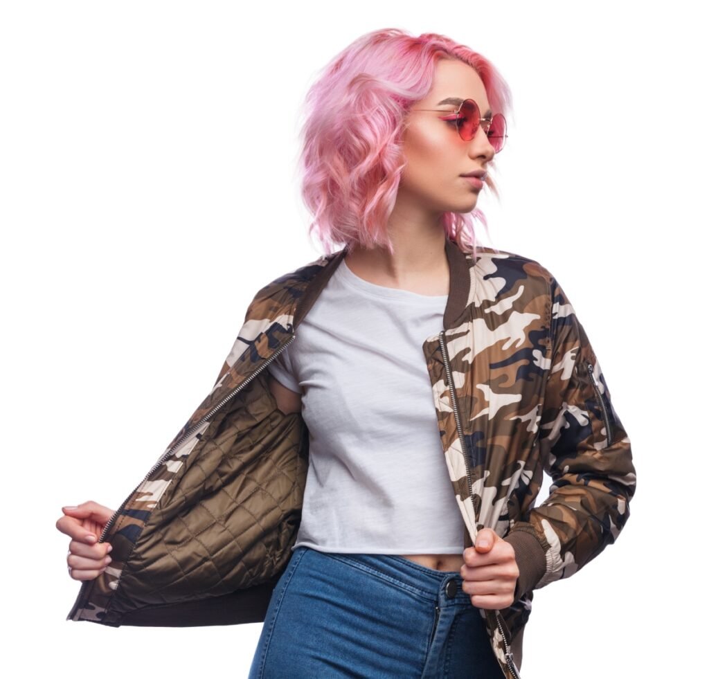 A stylish person with short pink hair and pink-tinted sunglasses wearing a camouflage nylon bomber jacket over a white crop top and blue jeans. They are turning to the side and pulling open the jacket to reveal the quilted inner lining.