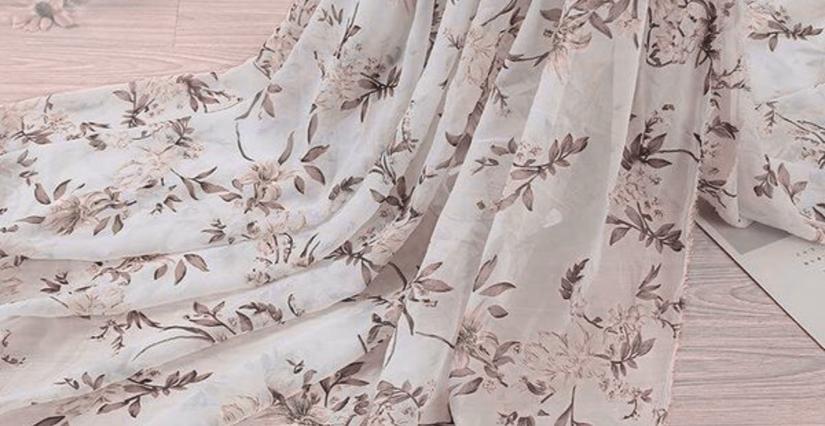 Soft, sheer organza fabric with a delicate floral print in light pink and beige tones draped over a pale wooden floor. Printed Organza Fabric
