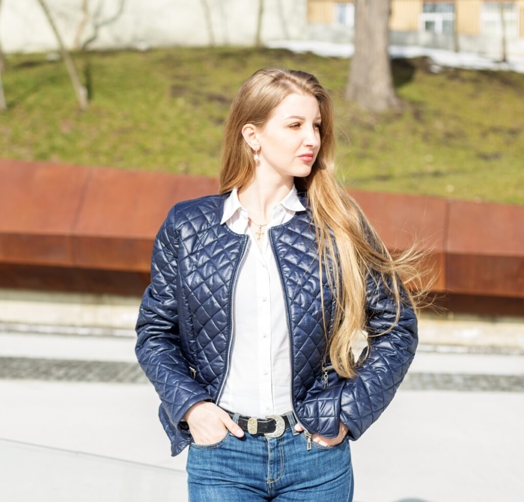 A stylish woman wearing a navy quilted bomber jacket over a white button-down shirt and blue jeans, standing outdoors with hands in pockets.