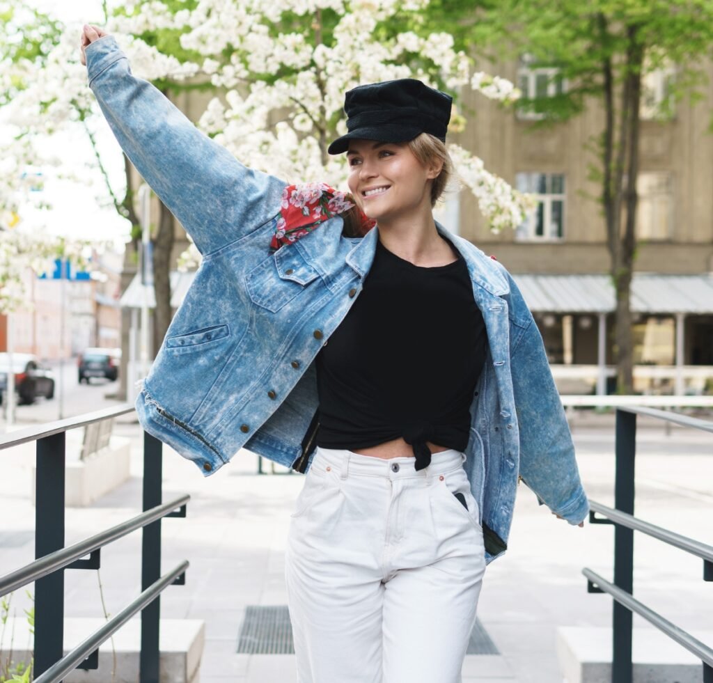 A stylish person wearing a light-washed denim bomber jacket over a black tied-front top and white jeans, posing outdoors with one arm raised in a playful stretch. They wear a black cap and a floral scarf peeking from the jacket, with a city street and blooming trees in the background.