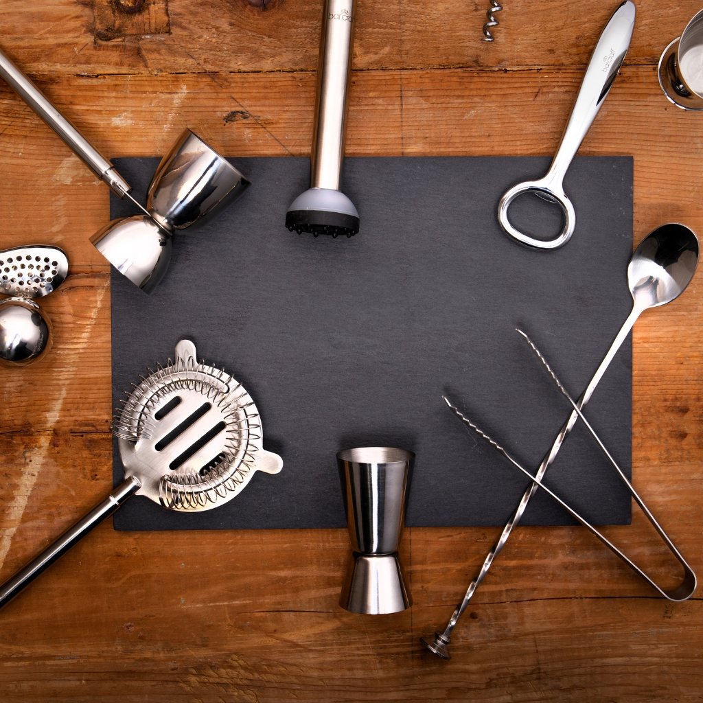 A collection of stainless steel bar tools arranged around a dark slate board on a wooden surface.
