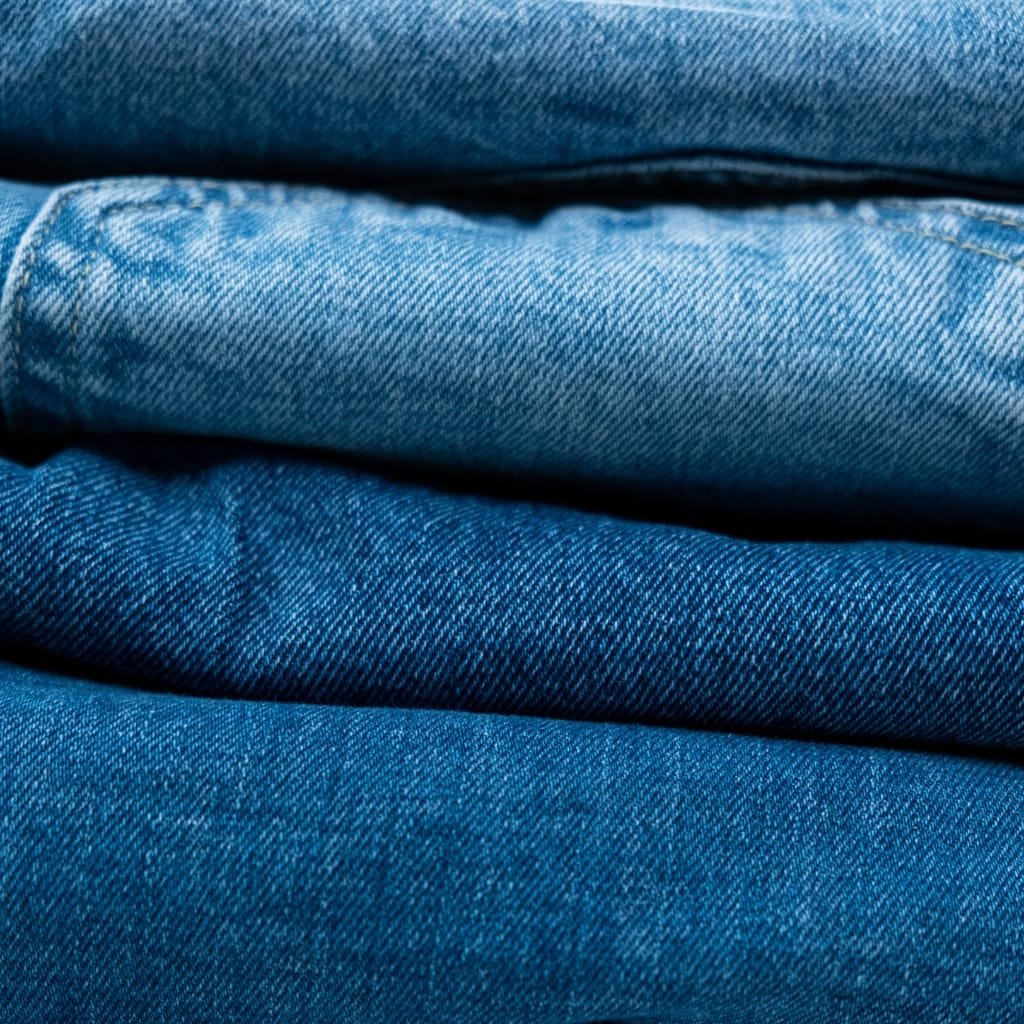 A close-up view of stacked folded blue denim fabric in varying shades, showing the diagonal twill weave and texture.