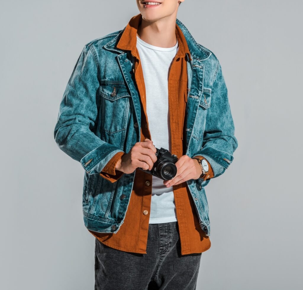 A person wearing a blue denim bomber jacket over an orange-brown overshirt and white tee, holding a small camera.