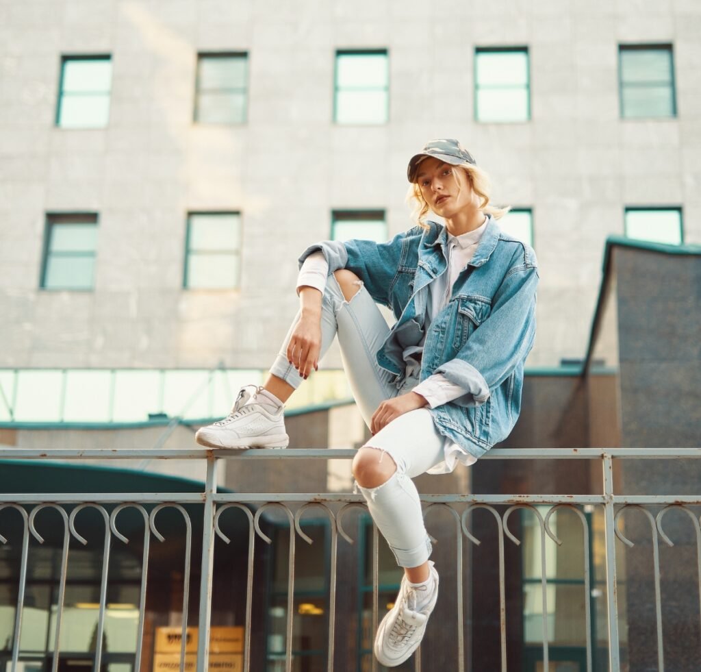 A stylish person sits on a metal railing outdoors in an urban setting, wearing a light-wash denim bomber jacket, ripped white jeans, white sneakers, and a cap.