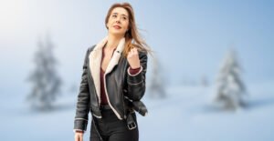 A woman wearing a black shearling bomber jacket with a white shearling collar stands outdoors in a snowy landscape.
