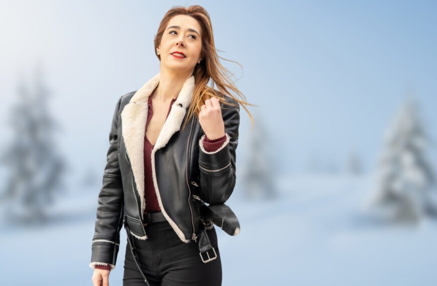 A woman wearing a black shearling bomber jacket with a white shearling collar stands outdoors in a snowy landscape.
