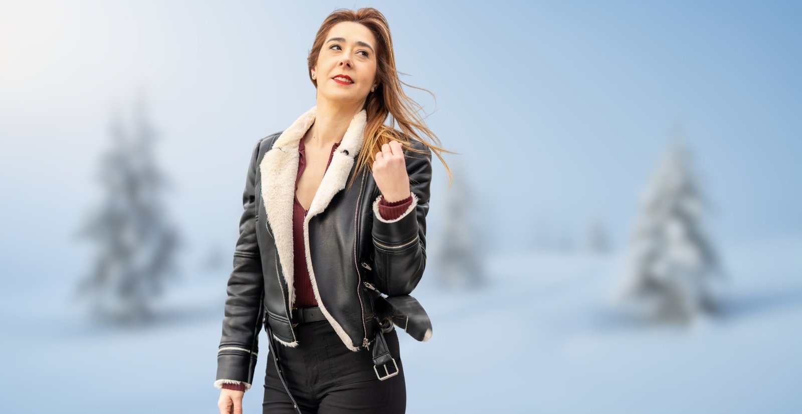 A woman wearing a black shearling bomber jacket with a white shearling collar stands outdoors in a snowy landscape.