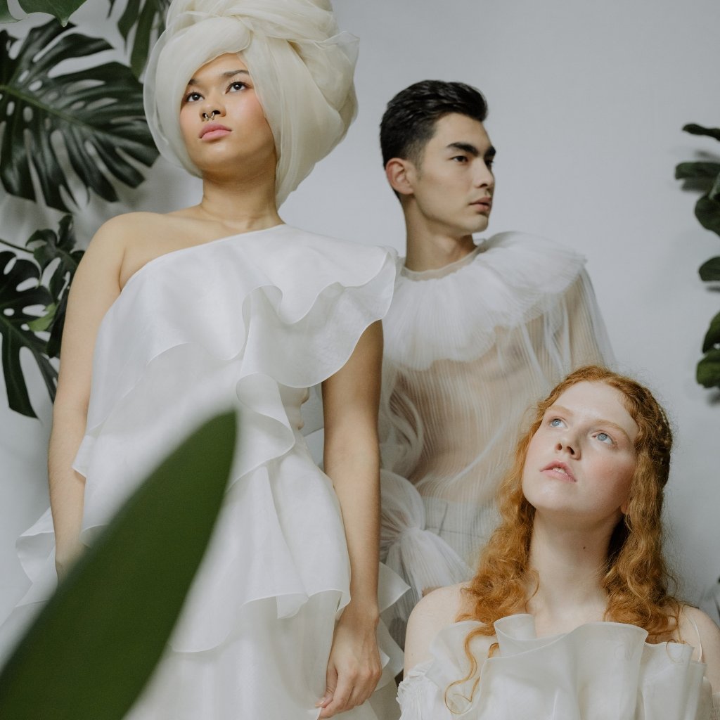 Three models wearing delicate sheer organza outfits: a woman in a white asymmetrical dress with ruffles and a large headwrap, a man in a translucent, pleated top, and a red-haired woman in a light, shimmery blouse. Surrounding greenery creates a soft, ethereal atmosphere