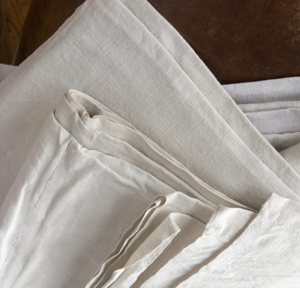 “White linen sheets and pillowcases neatly folded and layered, showing soft creases and a natural textile texture.”