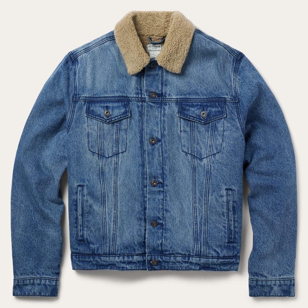 A blue denim jacket with a tan sherpa collar, button-front closure, two chest pockets with flaps, and vertical seam detailing, shown against a light background.