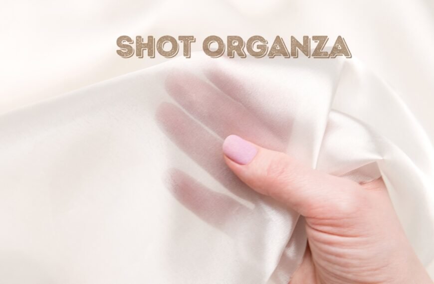 A close-up photo of a hand gently grasping a sheer, soft white organza fabric. The fabric folds create delicate shadows and highlights, and a light pink manicure is visible on the fingernails.
