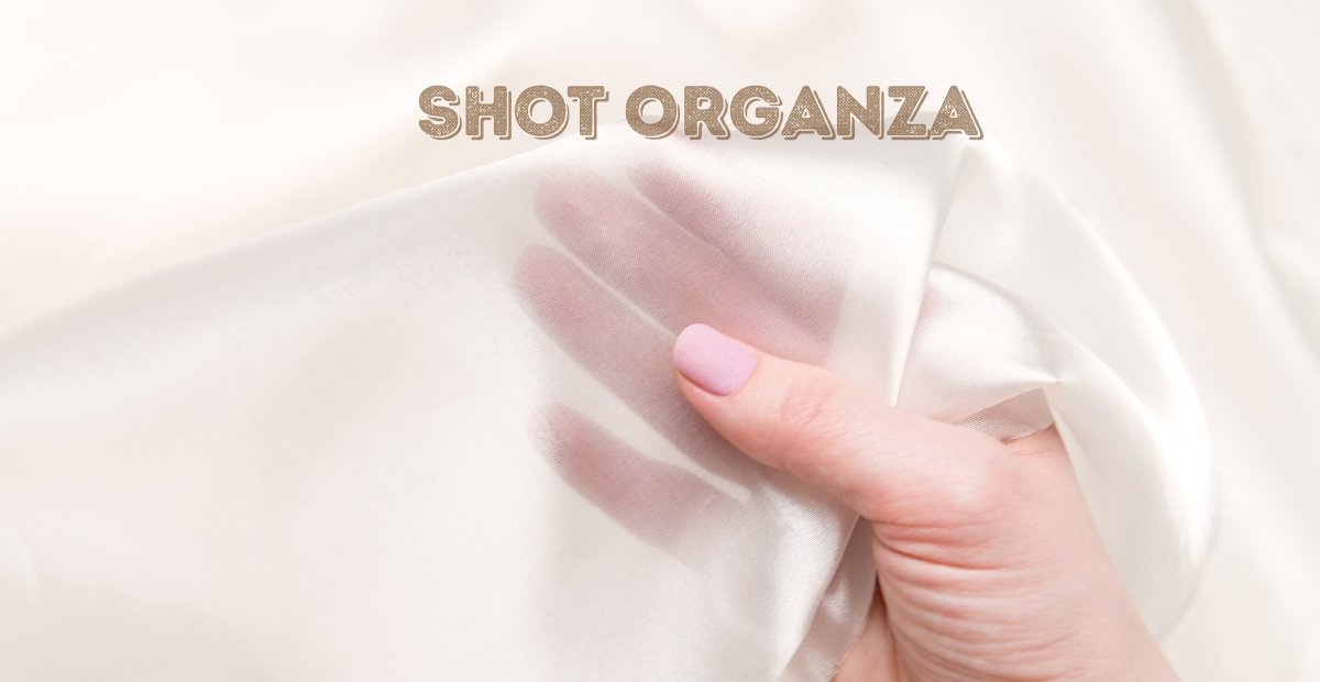 A close-up photo of a hand gently grasping a sheer, soft white organza fabric. The fabric folds create delicate shadows and highlights, and a light pink manicure is visible on the fingernails.