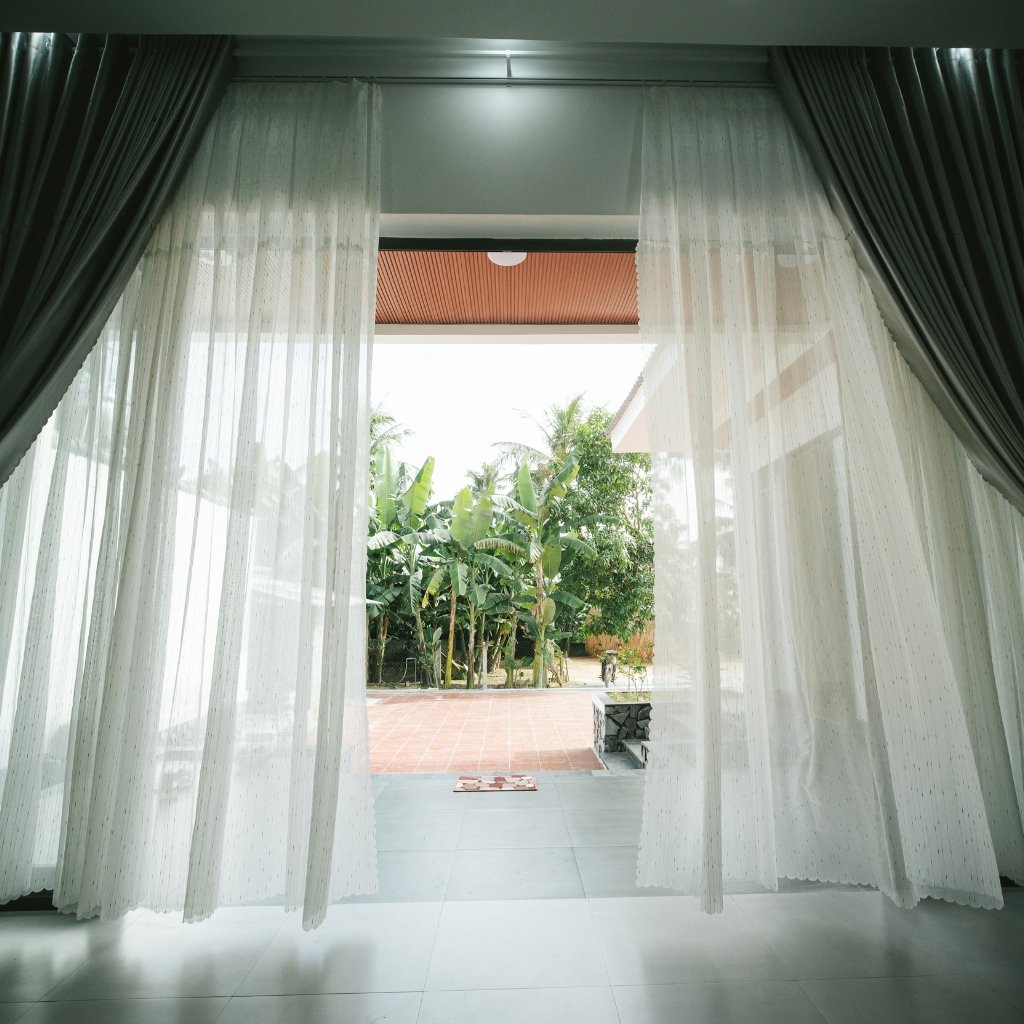 A bright room opening through sheer white organza curtains to an outdoor patio with lush green plants. The curtains frame a doorway, with tiled flooring inside and a brick-paved area outside. Soft daylight filters through the delicate fabric.