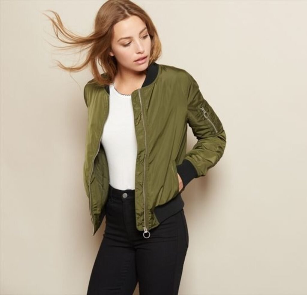 "A stylish olive-green bomber jacket with a black ribbed collar, wearing a white top and black jeans. The model has one hand in her pocket and her hair flowing slightly to the side."