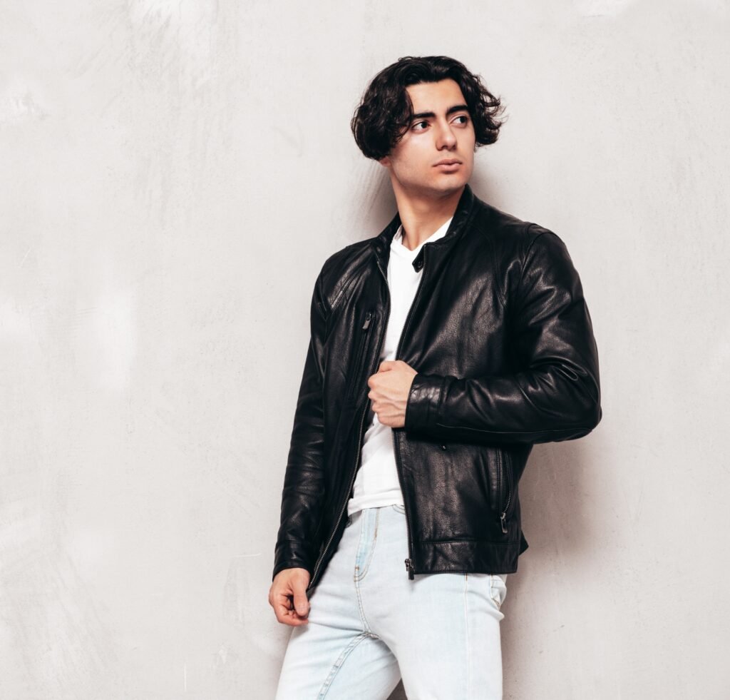 "Young man with wavy dark hair wearing a black leather slim-fit bomber jacket, white t-shirt, and light jeans, standing against a light gray textured wall."