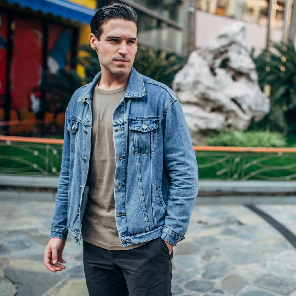 Man wearing a light-wash denim jacket over a beige t-shirt, walking outdoors in an urban street setting with trendy, casual streetwear style.