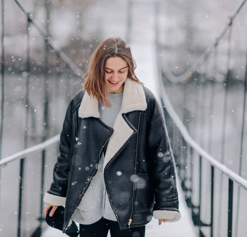 A smiling woman walks on a snow-covered pedestrian bridge wearing a black shearling bomber jacket with cream faux shearling lining visible at the cuffs, collar, and front edges. She pairs it with a light gray knit sweater and black pants. Snowflakes fall gently around her against a blurred, wintry background of railings and cables.