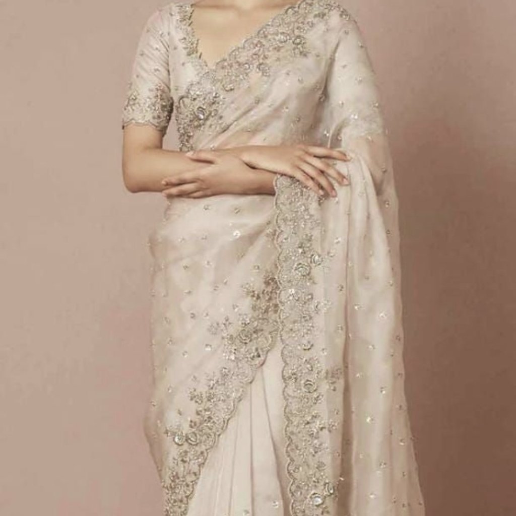 A woman wearing an elegant, ivory-beige saree with intricate silver embroidery and beadwork. The saree features a sheer, embellished border draped over the shoulder, short sleeves with lace-like embroidery, and scattered reflective embellishments across the fabric. The overall look is graceful and luxurious.