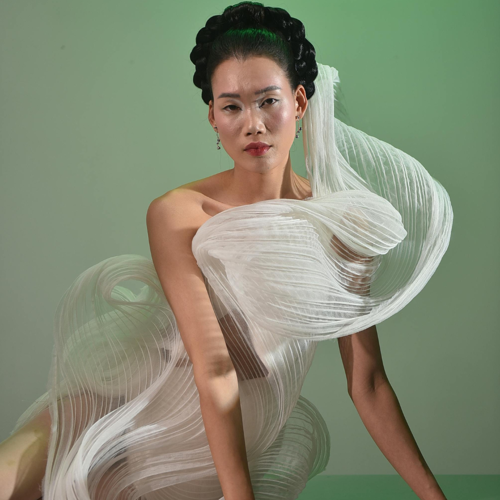 A fashion model wearing an ethereal, sculptural gown crafted from crushed organza. The fabric forms soft, airy waves around her body, with a sheer, lightweight texture. Her hair is styled in a dramatic braided updo, and she stands against a muted green backdrop, exuding elegant poise.